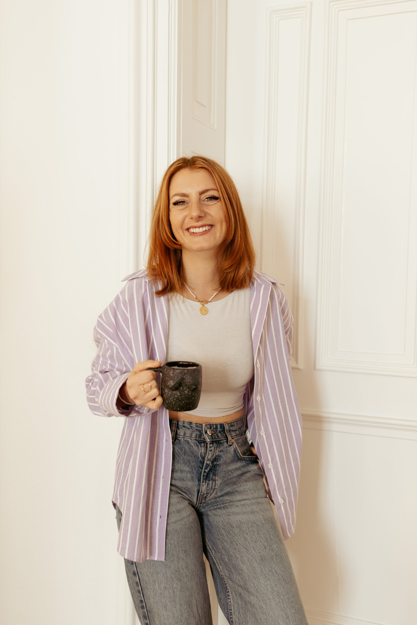 Mona CEO von einfachyou Werbeagentur lachend mit einer Tasse in der Hand, sehr sympathisch und aussagekräftig. Rotes Haar, weißer barocker Hintergrund, Weißes Shirt, Jeans und Lila Hemd mit Streifen.